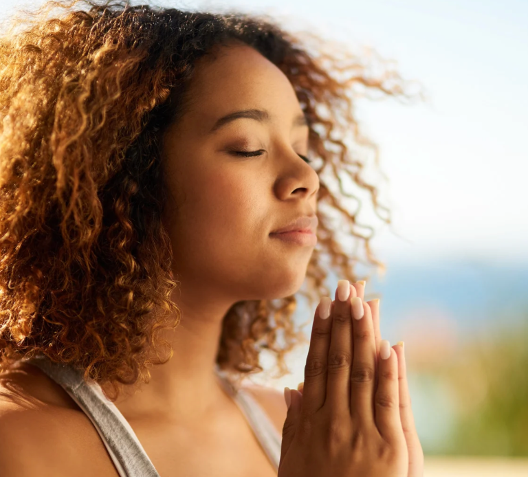 Young woman practicing mindfulness meditation outdoors with eyes closed and hands in prayer pose, enjoying peace and relaxation in natural sunlight.